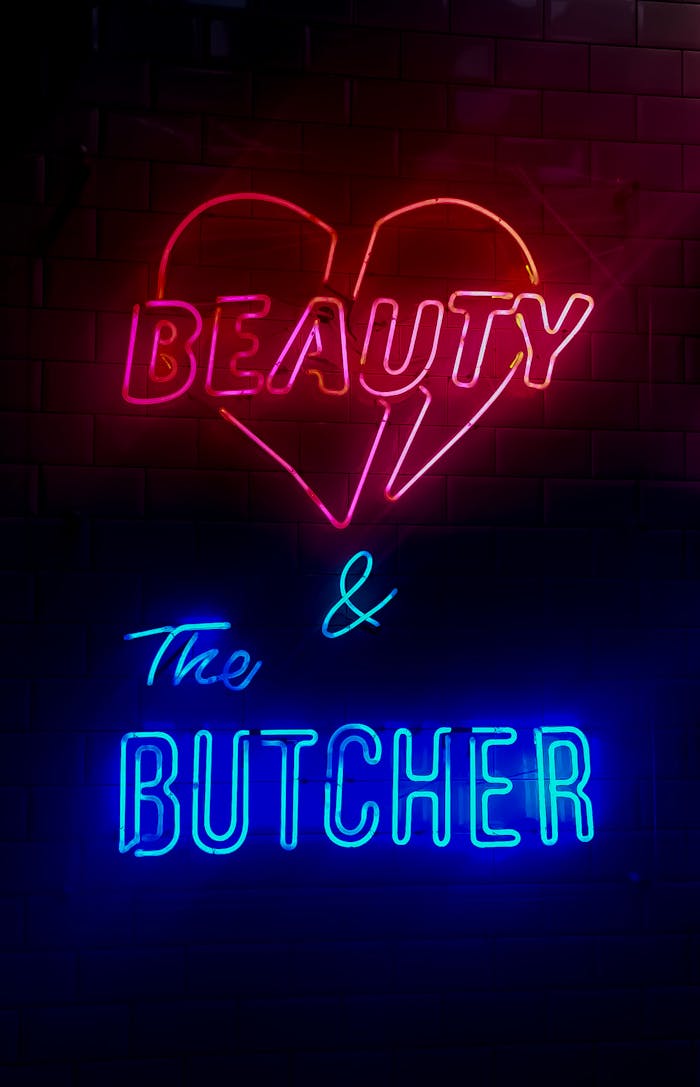 about-img Vibrant neon sign displaying 'Beauty & The Butcher' against a brick background, capturing urban nightlife ambiance.